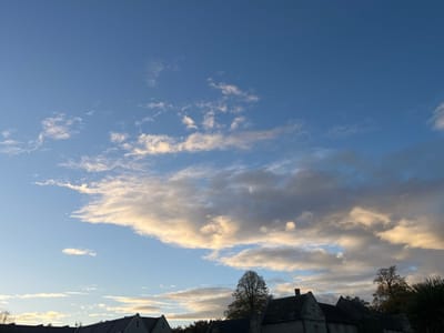The image displays an external view dominated by a dynamic sky over the village of Newton St. Loe, United Kingdom. The upper two-thirds of the frame consists of a deep blue sky transitioning to lighter hues towards the horizon. Scattered cirrus-like clouds are visible in the upper sections, while the middle and lower sky are filled with stratified clouds exhibiting varying tones of grey, white, and golden-yellow, suggesting low-angle sunlight, likely during sunrise or sunset. The bottom third of the image reveals the upper portions of architectural structures and deciduous trees. Multiple dark-grey pitched rooftops of stone or slate construction are visible, indicating traditional buildings. Several trees, some appearing bare or with sparse foliage consistent with autumn, are interspersed among the structures and along the horizon. No individuals, direct actions, or interactions are present in the static scene.