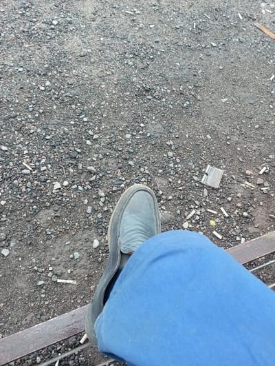 The image shows a person's left leg and foot, wearing blue pants and a grey shoe, resting on a metal beam. The ground is covered in gravel and dirt, with several cigarette butts and a piece of metal debris scattered around. The scene appears to be outdoors in an industrial or undeveloped area. The image does not provide any specific indicators to determine its location within the city of Бәйтерек, Kazakhstan.