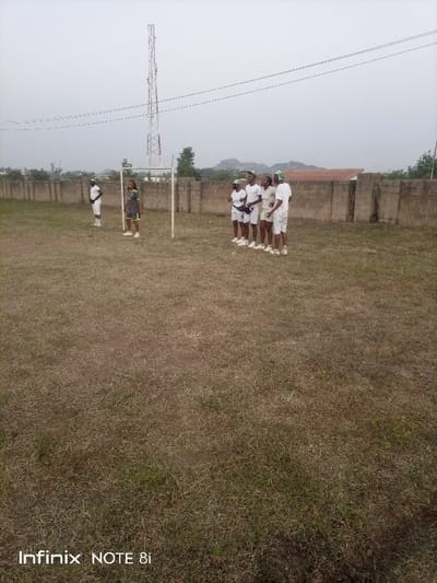 A group of seven people stand on a grassy field. Six are wearing white outfits, possibly uniforms, and stand in a line. One individual wears a darker outfit and stands a distance away near a goalpost. A tall communication tower is visible in the background, along with a wall bordering the field. The sky appears overcast. The scene suggests a training or practice session.