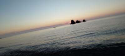 The image depicts a blurred seascape at dusk or dawn. A gradient of soft blues, pinks, and oranges colors the sky above a body of water with visible ripples. Silhouetted against the horizon are three dark, rocky formations protruding from the water. The foreground consists of a dark, indistinct area, possibly a shoreline or a boat. The motion blur suggests the photograph was taken while in motion, such as from a moving vehicle or vessel. The location is identified as Aleksandrovsk-Sakhalinskiy, Russia, which is a port city located on the northeastern coast of Sakhalin Island. This context implies the scene is likely the Sea of Okhotsk. There are no people visible in the image.