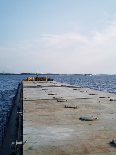 The image depicts a perspective view from the deck of a large barge moving across a body of water. The barge's deck is a wide, flat expanse of weathered concrete or metal, marked with numerous circular hatches. The water is a deep blue, with gentle ripples indicating calm conditions. In the distance, a tree-lined shore is visible, with a few structures and what appears to be a pylon on the left side. Further out on the water, a cargo ship is visible on the horizon. The sky is a pale blue with sparse, wispy clouds. The overall scene suggests a maritime or industrial setting, possibly in a large river or a lake, under a clear sky. The location is stated to be the city of Bessolovo, Russia.