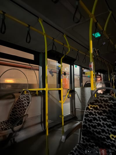 A dimly lit, canted perspective interior shot of a bus, possibly in Amadora, Portugal. Yellow handrails and straps are visible along the ceiling and sides. A woman wearing sunglasses is seated near the front of the bus. There's a seat with a black and white patterned cover. Outside the windows, indistinct urban elements are seen, with hints of buildings and street lighting. A green emergency exit sign is also visible. A bottle of "DuvVale" is partially visible on the seat.