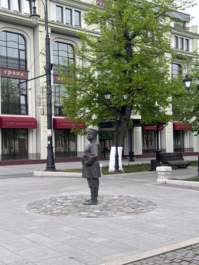 The image shows a statue of a man standing on a cobblestone mosaic embedded in a paved sidewalk. Behind the statue is a multi-story building with signs in Cyrillic script, indicating a location in a region with Russian-speaking influence. The building has dark-framed windows with metal railings and dark red awnings over the windows. A tree grows near the statue and partially obscures the view of the building. The image appears to be taken in Akhieli, Georgia, based on the Cyrillic signage.