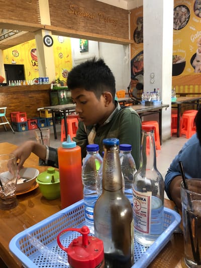 The image depicts a young boy in a green shirt sitting at a wooden table in what appears to be a restaurant. The background shows yellow walls with graphics, wooden beams, and posters. On the table, there are several bottles of drinks, including what looks like soy sauce and vinegar, a green bowl, and a red squeeze bottle with a blue cap. A blue plastic basket holds some of these items. The boy is looking down, seemingly engaged with the table. The overall setting suggests a casual dining environment. The presence of Indonesian text on the posters and labels indicates the location is in Indonesia. Uteungeulinggang is not a known city in Indonesia; it may be a village or a very small town.