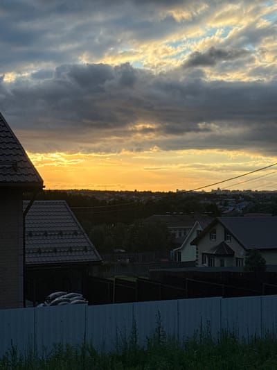 The image depicts a suburban landscape at dusk, with a dramatic sky featuring dark, heavy clouds interspersed with bands of bright orange and yellow light, indicating the setting sun. In the foreground, the dark, sloped roof of a house is visible on the left. Further back, several residential buildings with similar dark roofs are scattered amongst trees. A dark wooden fence runs horizontally across the middle ground, separating different properties. Beyond the houses and trees, a distant cityscape with several taller buildings is visible on the horizon. The overall scene is one of quiet evening in a residential area.