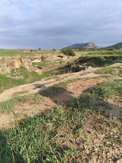 This is a landscape photograph taken in the city of Irgali, Russia. The image depicts a rural scene with rolling hills, a canyon or ravine, and distant mountains under a cloudy sky. The foreground is dominated by dry, sandy soil with sparse green grass and shadows. A pathway or track runs diagonally across the foreground. In the midground, a rocky canyon with steep, eroded walls is visible, covered in patches of green grass. Some trees are scattered across the landscape. In the background, larger hills and mountains are covered with vegetation, and a clear plateau is visible on the largest mountain peak. There are no people or animals visible in the image. The scene appears to be an outdoor natural environment, possibly with some evidence of historical structures or ruins within the canyon.
