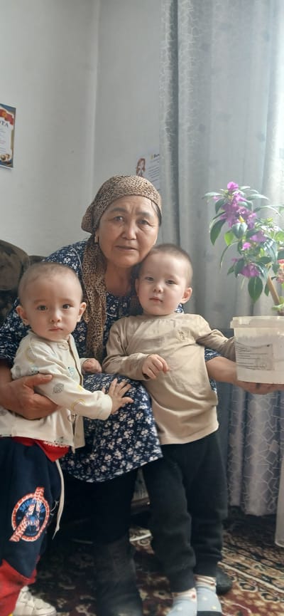 An older woman wearing a leopard print headscarf and a dark blue floral dress is seated and holding two young children. The child on her left is wearing a light-colored pajama top with cartoon images and dark blue pants with red accents and a Spider-Man logo. The child on her right is wearing a tan long-sleeved shirt and black pants. The woman is looking at the camera and smiling slightly. Behind them, a potted plant with purple flowers is visible next to a light-colored curtain with a geometric pattern. On the wall to the left, there is a certificate or award. The scene appears to be indoors, possibly in a living room, in the city of Mihaylovka, Kyrgyzstan.