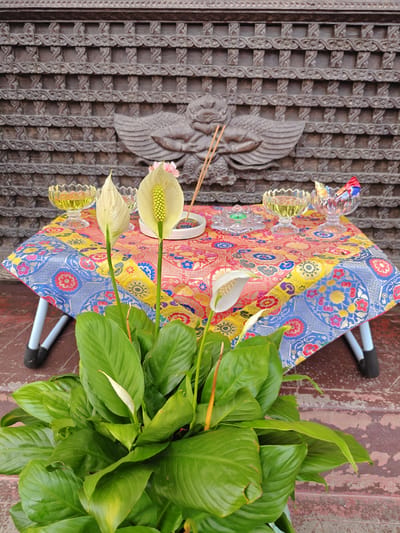 This is a medium shot, eye-level, outdoor photograph of a small table set for a ritual or offering. The table is covered with a colorful, patterned cloth featuring floral motifs in red, blue, yellow, and pink. On the table are several items: two clear glass bowls filled with a yellow liquid, a clear glass bowl containing wrapped candies, a small clear glass dish with a green object inside, and three incense sticks resting in a small white bowl. Two white Peace Lily flowers, each with a distinctive spadix, stand prominently in the foreground, with one in full bloom and the other partially open. Behind the table is a large, intricately carved wooden panel with a decorative, repeating pattern and a central relief carving of a winged mythical creature. The overall setting appears to be on a weathered, reddish-brown tiled surface, possibly a step or porch, with a slightly out-of-focus green plant in a pot in the lower foreground. The location is identified as Gokarneshwar, Nepal, suggesting a cultural or religious context for the scene. There are no people visible in the image.