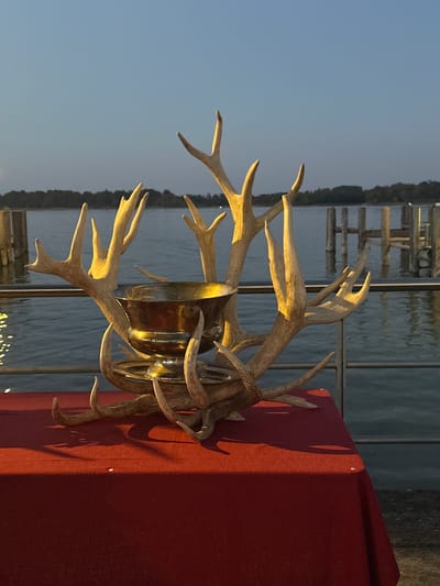 The image depicts a trophy made of antlers and a metal bowl, displayed on a red tablecloth. The trophy is placed on a table with a railing and a body of water in the background. Several wooden pilings are visible in the water, suggesting a waterfront location such as a pier or marina. The sky is clear and dusky, indicating either sunrise or sunset. The overall scene is an outdoor setting near a body of water, possibly in Prien am Chiemsee, Germany, given the context provided. There are no people visible in the image.