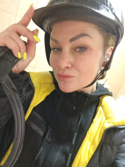 A close-up selfie taken within the city of Gorka, Russia, features a female subject in an indoor setting with light-colored, possibly tiled, walls. The subject has fair skin with visible freckles, blue eyes, and dark, well-defined eyebrows. She wears a black equestrian helmet with the brand "PONDHAUS" visible on the top, a black hooded sweatshirt with "ASTACEOO" visible on the drawstrings, and a black and yellow puffer jacket. Her left hand, with bright yellow nail polish, is positioned near the brim of her helmet. She maintains direct eye contact with the camera, exhibiting a subtle pout. A dark, padded strap, possibly from equipment or a bag, is partially visible on the lower left.