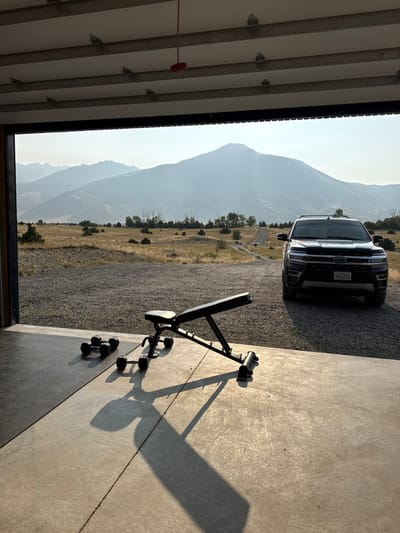 The scene depicts an open garage or structure in a rural area of Livingston, United States, offering a view of a mountainous landscape. In the foreground, on a concrete floor, a black adjustable weight bench is positioned with its seat angled left, casting a long shadow. To the left of the bench, two pairs of black hexagonal dumbbells rest on the concrete, also casting shadows. Overhead, a ceiling with parallel beams and a red pull cord is visible.
Beyond the concrete floor, a gravel expanse features a black Ford Expedition SUV parked on the right, facing slightly left. Its California license plate displays "9KVJ236." The midground consists of dry, golden-brown grassland with sparse green shrubs, and a dirt road curving into the distance on the left. The background is dominated by multiple layers of hazy, blue-grey mountains under a clear, bright sky. No individuals are present.