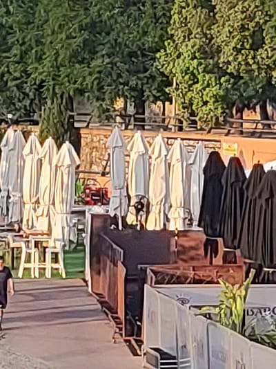 The image depicts an outdoor scene in Badajoz, Spain, likely a patio area of a restaurant or establishment. In the foreground, a person wearing a black t-shirt is walking away from the camera. Behind them, several white umbrellas are closed and lined up, some with ruffled edges. To the right, there are several black umbrellas, also closed. In the background, trees with dense green foliage are visible. There are also glimpses of tables and chairs, suggesting a seating area. The overall impression is a quiet, possibly early morning or late afternoon scene, with the umbrellas indicating a space designed for outdoor dining or relaxation. The lighting suggests it's daytime, with sunlight casting shadows.