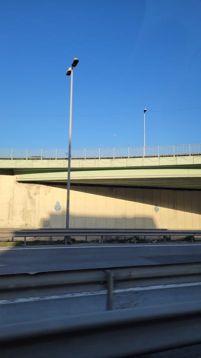 The image shows an underpass or overpass structure in Beylikdüzü, Turkey, under a bright blue sky. Two streetlights are visible against the sky. The structure is painted in light green and beige. A decorative logo is painted on the beige wall. The road is beside the structure, and metal guardrails run along the edge of the road. A car is visible in the foreground.
