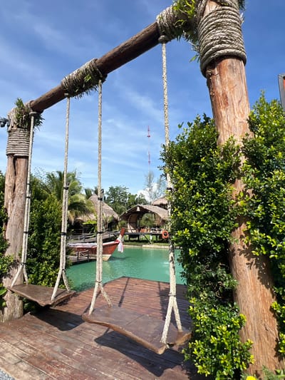 This image depicts a rustic outdoor scene with two wooden swings suspended from a sturdy wooden beam, supported by thick wooden posts wrapped in rope. The swings hang over a wooden deck, which leads to a tranquil body of turquoise water. In the background, traditional thatched-roof huts and palm trees suggest a tropical setting. A longtail boat is partially visible on the water, adding to the local ambiance. The scene is set against a bright blue sky with scattered white clouds. The overall impression is one of a peaceful, resort-like environment, likely in a location like Tambon Pa Kae Bo Hin, Thailand, known for its scenic beauty and relaxed atmosphere.