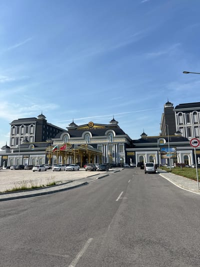 This image captures the exterior of the "GOLDENEYE CASINO" complex in Kapitan Andreevo, Bulgaria, under a clear blue sky with sparse, wispy clouds. The casino building is a large, ornate structure featuring a neoclassical architectural style with dark gray/black facades accented with gold trim and white columns. Multiple dome-like structures, some topped with spires, are visible across the roofline. The central section of the building prominently displays the "GOLDENEYE CASINO" logo in gold above a grand entrance with a golden arched canopy. Several classical statues in white are positioned near the entrance and along the building's facade.
In front of the main entrance, two flags are displayed: the national flag of Bulgaria and the national flag of Turkey. A banner on the right side of the complex advertises "YAZ AYRICALIĞI" (Summer Privilege) with "25€" and an image of a pool.
The foreground features a paved asphalt road leading directly towards the casino entrance, marked with white dashed lines. A white delivery van is visible on this road, facing the building. To the left of the road is an unpaved gravel parking area occupied by multiple parked cars of various makes and colors, predominantly white, silver, and dark. To the right of the road, a sidewalk and a strip of manicured grass with low vegetation are present. A lamppost stands on the right, and a red circular road sign with a white truck symbol and a diagonal line, indicating "no trucks" or "trucks prohibited," is clearly visible. Another section of the building, similar in architectural style, extends to the far right. The overall scene appears well-maintained and devoid of any prominent human figures.