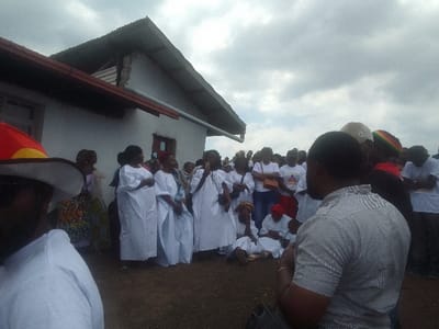 A gathering of people outside a white building under a cloudy sky. Several women stand together, wearing white robes, and one appears to be speaking into a microphone. Other attendees wear white shirts and various hats. One man in the foreground wears a white shirt and a wide-brimmed hat. The ground appears to be dirt or gravel. The group size suggests a ceremony or community event.