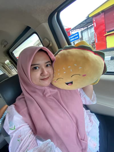 A woman wearing a pink hijab and a patterned pink dress is lying down inside a car. She rests her head on a plush, burger-shaped pillow with a smiling face. The car's sunroof is open, revealing a building with red and white elements and a sign that says "Radiator". The scene appears relaxed and comfortable, with a focus on the woman's serene expression. The lighting suggests it is daytime.