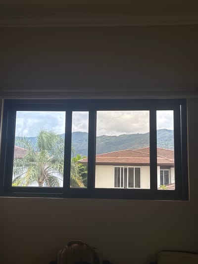 The image shows a view from a window in a house in Río Oro, Costa Rica, located at latitude 9.945 and longitude -84.201. The window is a sliding glass window with a black frame. It is divided into three panes. The leftmost pane shows a palm tree and a green bush, while the middle pane reveals a view of a mountain range. The rightmost pane displays the back of a white house with a brown roof. The house is likely located in the same neighborhood as the one the window is in. The scene is bathed in soft, natural light, and the sky is overcast. The windows and the house are clean and well-maintained. The scene is peaceful and tranquil.