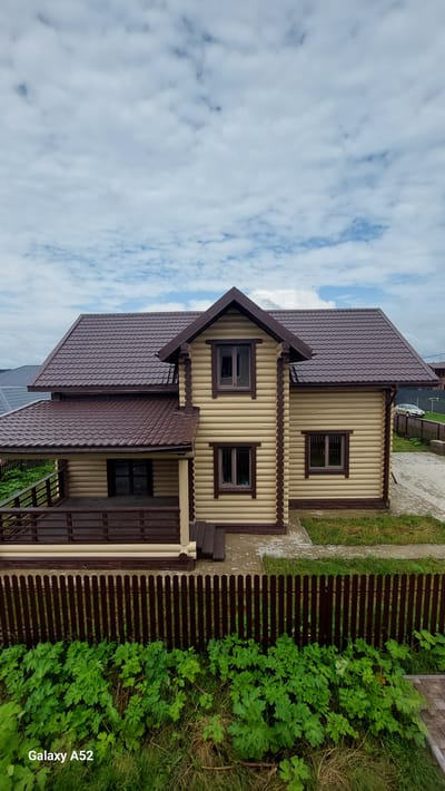 The image shows an aerial view of a two-story wooden log house with a brown metal roof, located in the village of Dolce Vita KP, Russia. The house features a porch with a dark brown railing. In the foreground, a dark brown wooden fence separates the property from lush green vegetation and grass. The background includes a paved driveway, a white car, and other houses with similar roofs. The sky is partially cloudy.