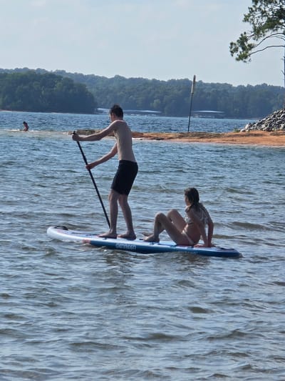 Two individuals are on a paddleboard in the water. A young male, standing, holds a paddle. A young female is seated on the paddleboard. The paddleboard is blue and white with "GENREEN" visible on its side. In the background, a sandy beach with rocks and trees lines the shore. A distant tree with green foliage stands on the right side of the frame. The sky is clear and blue. The location is Lake Lanier in Flowery Branch, Georgia.
