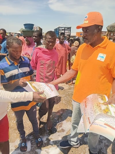 The scene takes place in Katabu, Nigeria (latitude: 10.707, longitude: 7.514). An older man, wearing a bright orange shirt with the word "KAMA" on it, is standing and handing a white flyer with the words "NATIONAL BUREAU" on it to a younger man standing in front of him. The younger man, wearing a navy blue and white striped shirt, appears to be reviewing the flyer as he holds it in both hands. Behind them, several other people stand in a crowded area, mostly men, all in different attire. The man in orange is wearing a dark blue jean and white sneakers. The younger man is wearing blue jeans with black athletic shoes. The scene takes place on a paved surface. The man in orange has a dark complexion and a thick beard, while the younger man has a light complexion. The background includes a truck, and a few other people are visible. The ground is covered in a light layer of water and a light dust. A man with a bucket on his head is standing behind the man in orange. The scene is brightly lit, indicating daytime.