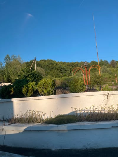 The image depicts an outdoor scene under a clear blue sky, possibly in Halych, Ukraine. In the foreground, there is a white concrete structure, possibly a wall or planter box, with some vegetation growing on top, including small plants with yellow flowers. The structure shows signs of wear and tear, with cracks visible. Behind the concrete structure, there are several green bushes and trees, forming a dense background. To the right, a tall, thin, orange pole rises into the sky. In the middle ground, an orange, abstract metal sculpture with arched elements and a light fixture is visible. Further back, a line of trees covers a hillside, with a power line supported by wooden poles visible among the trees. The sunlight suggests it is daytime, with some shadows cast on the concrete structure.