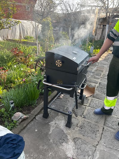 The image depicts a person operating a black charcoal grill in an outdoor setting. The person is wearing green and yellow work pants and blue rubber boots. Smoke is emanating from the grill. The grill is placed on a stone-paved surface next to a garden with flowers and greenery. In the background, there is a wooden structure and a brick building. There also appears to be a turtle statue next to the grill. This location may be in Bukovlak, Bulgaria.