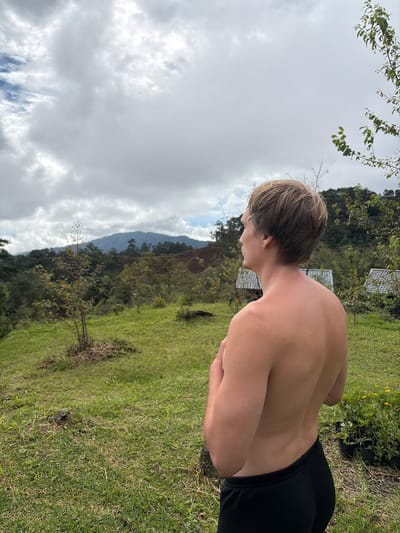 A shirtless young man with blond hair stands in a grassy area, facing away from the camera and looking towards a mountainous landscape under a cloudy sky. He is wearing black pants. The environment appears to be rural, with trees, some small structures with white roofs visible in the background, and lush green vegetation. The scene suggests a natural setting, possibly a park or a rural dwelling, in Ban Khun Wang, Thailand.
