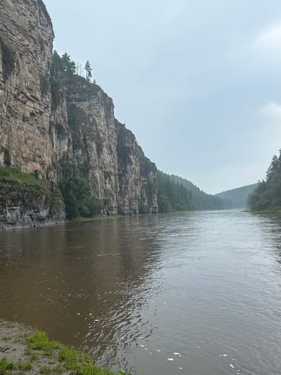 The image depicts a wide, murky river flowing between steep, rocky cliffs on the left and densely forested hills on the right. The sky is overcast and appears pale. The cliffs on the left are sheer and striated, with some sparse vegetation clinging to their surfaces. The river's surface shows ripples and some scattered white foam. The bank in the foreground on the left is muddy with patches of green grass. The forested hills in the distance are shrouded in a soft haze, and the overall impression is one of a rugged, natural landscape under a subdued sky. The location is identified as the city of Kul'metovo, Russia.