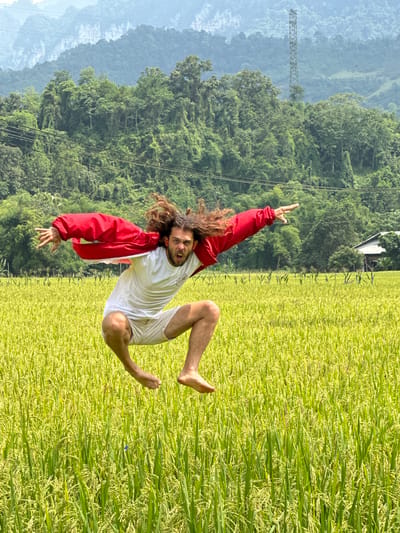 The image, correctly oriented, depicts a man joyfully leaping high above a vibrant rice paddy field under bright daylight. The man, with long, curly brown hair and a beard, is captured mid-air, wearing a white t-shirt, white shorts, and an open red jacket. He is barefoot, and his expression is one of exhilaration, with an open mouth suggesting a shout or laugh. His left hand is raised in a "rock on" gesture, while his right hand is splayed open below him.
The setting is a picturesque rural landscape in Ngọc Đường Commune, Vietnam. The foreground and midground are dominated by a lush field of rice, showing varying shades of green and yellow, indicating mature grains ready for harvest. In the background, rolling mountains covered in dense green foliage stretch across the horizon, appearing hazy in the distance. A tall metal transmission tower is visible on one of the distant mountain slopes, and overhead power lines traverse the sky above the tree line. The clear sky and strong illumination suggest it is daytime, likely mid-morning or afternoon. The overall mood conveyed is one of freedom, energy, and connection with nature. There is no visible text.