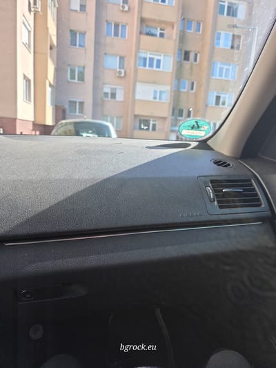 The image is taken from inside a car, likely parked in Krumovgrad, Bulgaria, given the "bgrock.eu" watermark. The view through the front passenger window reveals a multi-story building with numerous windows, some of which have air conditioning units installed. A green road tax sticker is visible on the inside of the windshield. Inside the car, the dashboard is visible, featuring an air vent and the word "AIRBAG" embossed on the panel.
