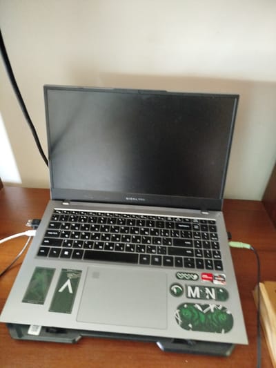 The image shows a laptop computer placed on a wooden desk. The laptop is open and appears to be resting on a cooling pad. The laptop has a silver keyboard and a black screen. There are several stickers on the palm rest of the laptop, including one with the letters "MN" and another with what appears to be a green design with two figures. A black cable runs from the top left corner of the laptop, and a green cable is plugged into the right side of the laptop. A portion of a wooden object, possibly a book, is visible on the right side of the desk. The background appears to be a plain wall. The image is taken from a slightly elevated angle, looking down at the laptop. The location of Зиянчурино, Russia is not depicted or suggested by any elements within the image.