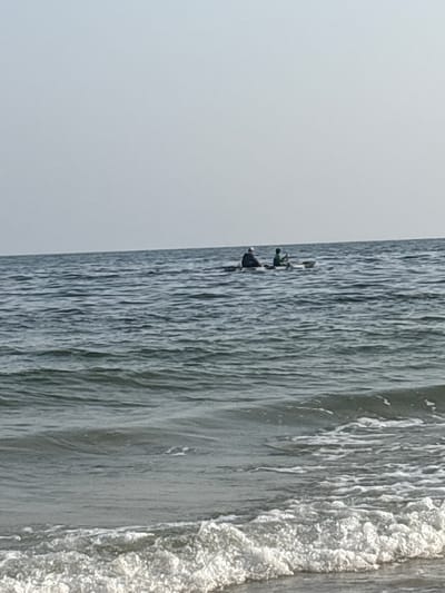 The image depicts a serene view of the ocean, with two individuals in a small boat or kayak approximately in the middle distance. The boat appears to be a light color, possibly white or beige, and it is moving across the water. One individual, wearing a hat, is positioned towards the front of the boat, and another individual, wearing a green shirt, is seated behind them. Both individuals seem to be engaged in a water-based activity, possibly fishing or paddling, as one appears to have a fishing rod extended. The water is a deep blue-gray color with gentle ripples and some foamy whitecaps in the foreground, suggesting a calm sea with a slight breeze. The horizon line is clearly visible, separating the water from an overcast sky that is a pale, uniform gray. The image gives the impression of a peaceful day at sea, possibly on the coastline of Ajman, United Arab Emirates, known for its beautiful beaches and calm waters.