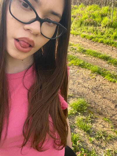 A selfie shows a young woman with brown hair wearing black-rimmed glasses and a pink shirt. Her tongue is slightly out. The background consists of a dirt and grass area. No information indicates a location in Blaskovo, Bulgaria.