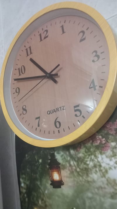 This is a close-up, eye-level shot of a round, yellow-framed clock with a light brown face, hanging on a wall. The clock has black hour, minute, and second hands, and black numerals indicating the hours, from 1 to 12. The word "QUARTZ" is printed on the clock face. The clock is slightly tilted. The background behind the clock is a blurry image of a nature scene, featuring trees, pink flowers, and a lit lantern hanging from a branch. The image does not contain any people or indicate a specific location within Oliveira do Bairro, Portugal.