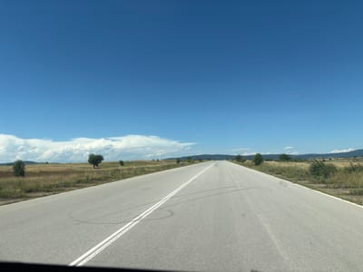 The image shows a straight, empty asphalt road extending into the distance under a clear blue sky with scattered white clouds. The road has a double white line marking its center. On either side of the road is dry, grassy terrain with scattered trees. In the distance, rolling hills are visible. There are no people or vehicles visible in the frame. The scene appears to be a rural highway, likely in a temperate climate during the daytime. The location is specified as Mladinovo, Bulgaria.