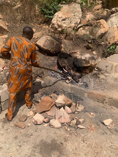 In Egbedi, Nigeria, an adult male stands with his back to the viewer, facing a functional fire pit or forge constructed amidst natural rock formations and concrete blocks. He is dressed in a full-length, two-piece orange-brown garment featuring a black and white leaf pattern, paired with light brown sandals. The man uses a long, dark metal rod to manipulate glowing embers and a visible reddish-orange object within the active fire. The immediate ground is composed of dry, light-colored soil, with a cluster of various sized rocks and broken masonry fragments in the foreground. The background features large, weathered rocks and sparse green vegetation under direct sunlight.