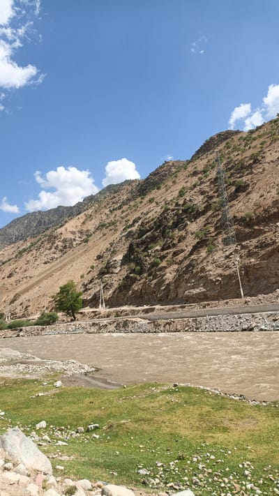 A wide, muddy river flows through a valley, with a steep, rocky mountainside rising on the right. The mountainside is sparsely covered with vegetation and features a power line tower. A road runs along the base of the mountain, parallel to the river. The foreground on the left consists of a grassy embankment dotted with rocks. The sky is blue with scattered white clouds. The image appears to be taken in a natural, rugged landscape, likely a rural area outside of a city.