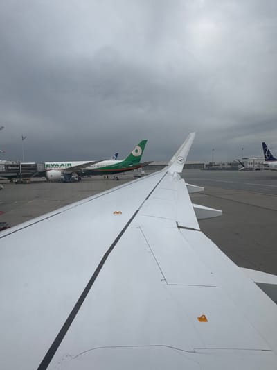 The image displays an airport apron scene under an overcast grey sky at Munich Airport (MUC) in Oberding, Germany. In the foreground, the clean white wing of a passenger aircraft, featuring a partial Lufthansa logo on its wingtip, extends from the left. This wing has a prominent black longitudinal stripe and two visible orange tie-down rings.
In the mid-ground, a white EVA Air passenger aircraft, identifiable by "EVA AIR" on its fuselage and a green tail with an orange globe logo, is parked at a gate. It is connected to a jet bridge that has "Revolut" branding near its attachment point. Several pieces of ground support equipment, including baggage carts and a blue ground power unit, are positioned around the EVA Air aircraft's underside. Multiple individuals, appearing to be airport ground staff, are visible on the tarmac near the aircraft's fuselage. Partially visible behind the EVA Air aircraft is the blue tail of an ANA (All Nippon Airways) aircraft.
Further in the background, another stationary passenger aircraft with a blue tail featuring a white, stylized bird logo, identifiable as LOT Polish Airlines, is visible on the tarmac. Airport terminal structures are discernible in the distance. The scene indicates ongoing airport ground operations.