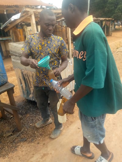 Two adult males are engaged in transferring liquid from a larger container to smaller bottles using a funnel and a water bottle. The scene appears to be outdoors, likely in a market or roadside stall environment in Madakiya, Nigeria, indicated by the rustic wooden structures and uneven ground with gravel and dirt.
The man on the left, wearing a blue and yellow patterned short-sleeved shirt and dark trousers, is holding a green funnel over a brown bottle. He is looking down at his task. To his right, the second man, dressed in a teal green t-shirt with orange lettering and light denim shorts, is holding a partially filled plastic water bottle, seemingly assisting in the transfer process. His body is angled towards the first man, and he is also looking down.
Various objects are visible in the background and foreground. To the left, a large blue plastic container is partially visible. To the right of the man on the left, a series of brown bottles and a lighter colored bottle are present. The ground is covered with small stones and dirt. Behind the men, a simple structure with a corrugated metal roof and wooden supports is present, suggesting a rudimentary stall or shelter. In the further background, other figures and stalls are discernible, hinting at a populated area. The overall lighting suggests it is daytime.
