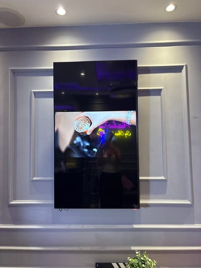In Sweileh, Jordan, a vertical, black, rectangular screen is mounted on a light purple, paneled wall. The screen displays a video feed showing a person's hand holding a clear glass filled with brown material. Below the hand, a neon sign with yellow and pink lettering is visible, with a blue neon camera icon to the left. A reflection on the screen shows a person wearing a sparkly silver top and black pants, holding a phone. The wall features decorative molding, and a white circular speaker is installed on the ceiling above. The lighting on the wall is soft, with a focused spotlight from the ceiling casting a shadow on the molding.