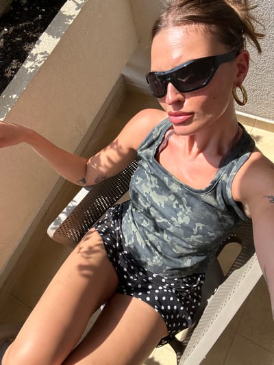 An adult female is observed seated in a light-colored, perforated chaise lounge on an outdoor balcony or terrace in Zadar, Croatia. She is positioned facing slightly right of the viewer, with her left arm extended horizontally. She wears a dark green camouflage-patterned tank top, black shorts with white polka dots, and large black wrap-around sunglasses. A prominent gold hoop earring is visible on her right ear. Tattoos are present on her left forearm and potentially her right arm. Strong, direct sunlight illuminates the scene, casting distinct shadows of foliage onto her body and the light-colored stucco wall behind her. A dark planter or soil bed is partially visible in the upper left background.