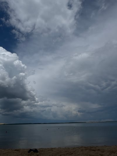 A frontal, eye-level shot captures a serene body of water, possibly Lake Aiyrtau, in Kazakhstan, under a dramatic, cloudy sky. The water stretches to the horizon, with a line of distant trees and faint hills visible. The foreground is a sandy beach, with a pair of dark flip-flops resting on the sand. Several buoys, red and white, are scattered across the water's surface, suggesting a designated swimming or boating area. The sky is a dominant feature, filled with a mix of dark, heavy storm clouds and brighter, white cumulus formations, hinting at impending or recent rainfall. The overall mood is somber and tranquil, with a sense of nature's power and vastness.