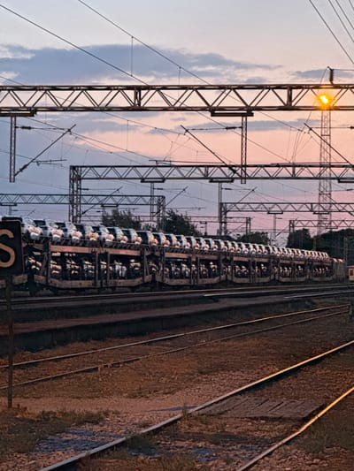 A low-angle, rotated image shows a train on tracks in Ruma Municipality, Serbia. A metal electrical pole structure with a lit light fixture stands prominently to the left. Overhead electrical lines crisscross against a dusky sky of pinks and greys. The train cars are stacked high with a repeated pattern of containers, possibly vehicles. The tracks run parallel to the right side of the frame, with earth and vegetation bordering them.