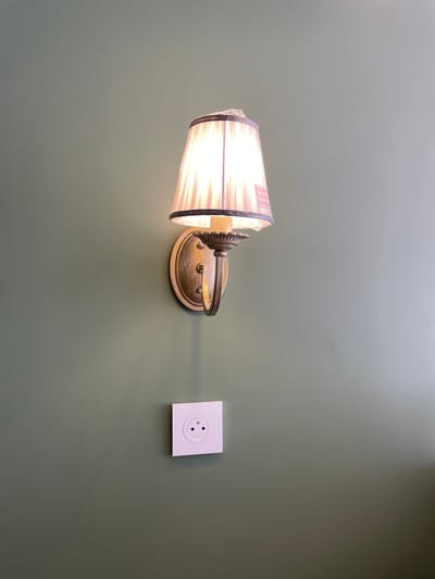 A wall-mounted lamp with a pleated white shade casts a warm glow on a textured, green wall. Below the lamp, a white electrical outlet with two round openings is visible. The lamp features a brass-colored base and arm. The overall scene suggests an indoor setting, possibly a hotel room or a hallway. There is no indication of the specific location in the city of Meaux, France.
