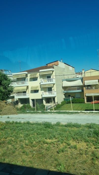 The image depicts a residential building complex in Larissa, Greece. The building is cream-colored with multiple balconies adorned with white railings and yellow awnings. The windows are dark, and some have closed blinds. The building is situated in a suburban or semi-urban environment, with a clear blue sky overhead. In the foreground, there is a patch of dry grass and some green vegetation, suggesting an outdoor area. The overall scene portrays a typical residential neighborhood.