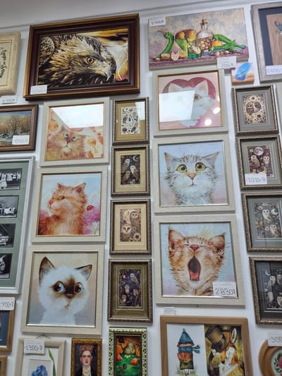 The image shows a wall in what appears to be an art gallery or shop, displaying a variety of framed artworks. The artworks are primarily paintings and prints, featuring subjects such as animals (cats, an eagle, owls, a fox), still life, and a portrait of Frida Kahlo. Some frames are ornate, while others are simple. Price tags are visible on several of the artworks, indicated by handwritten numbers followed by "P" (likely indicating Russian Rubles).

The specific location is identified as Arseniev, Russia, based on information provided separately, though there are no overt visual cues in the image itself to confirm this location.

Detailed breakdown of visible art pieces and prices:

*   **Top left:** A large framed artwork of an eagle's head, with a price tag of "10500 P". To its left, a partially visible framed artwork with a price tag of "6 000 P".
*   **Middle left:** A framed artwork of a snowy landscape with trees, priced at "850 P". Below this, a collection of smaller framed black and white photographs, one with a price tag of "700 P".
*   **Center, top:** A framed artwork of a white cat with large eyes inside a heart shape.
*   **Center, middle:** A framed painting of a fluffy orange cat with green eyes.
*   **Center, bottom:** A framed painting of a yawning orange cat. This has a price tag of "2830 P".
*   **Center, bottom right:** A framed artwork depicting a person dressed as a fox wearing a top hat next to a figure in a hat and scarf holding a pumpkin.
*   **Top right:** A still life painting of fruits and vegetables with a bottle, priced at "5 500 P". To its right, a small painting of a dolphin and a wave, priced at "700 P".
*   **Right side, stacked artworks:** Several framed artworks featuring owls in various poses and settings, some with moons and floral elements. One of these has a price tag of "3210 P". Another has a price tag of "300".
*   **Bottom center:** A framed portrait of Frida Kahlo. Below this, a framed artwork with a blue iris.
*   **Bottom right:** A framed artwork of a cat sitting on a table with apples.

The overall impression is of a densely packed display of art for sale. The lighting is even, suggesting indoor illumination. There are no people visible in the image.