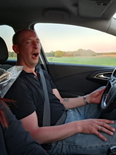 A man is seated in the driver's seat of a car, wearing a black shirt and light blue jeans. He is also wearing a seatbelt. His mouth is open, possibly talking or yawning. He has a wedding ring on his left ring finger and a bracelet on his left wrist. A tattoo is visible on his left forearm. The car's interior is visible, including the steering wheel and dashboard. Outside the window, there is a field, some trees, and a light-colored sky. The location appears to be the city of Любанскі сельскі Савет, Belarus, judging by the landscape and sky visible through the car window.