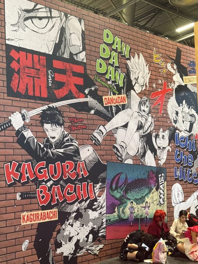 The image shows a large mural painted on a brick wall at Paris-Nord Villepinte Exhibition Centre in Villepinte, France. The mural features various manga-style illustrations, including characters with spiky hair, dramatic eyes, and action poses, along with Japanese text. Prominent titles visible are "KAGURA BACHI" and "DAN DA DAN". In the foreground, several young women are sitting on the floor, some looking at their phones. They appear to be attending an event or convention, possibly related to anime or manga given the artwork. One woman has bright red hair and is wearing a dark red jacket. Another is wearing a white shirt. The lighting suggests an indoor venue, likely a convention hall.