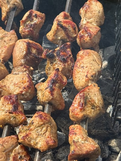 The image shows several skewers of meat being grilled over hot coals. The meat is cut into chunks and appears to be marinated, with visible herbs and spices on its surface. Some pieces are browned and slightly charred, indicating they are cooking well. The skewers are arranged on a metal grill over a bed of dark, glowing coals, which emit a faint haze of smoke. The background consists of more coals and parts of the grill grate. The lighting suggests it might be daytime, possibly outdoors given the barbecue setting. There are no people visible in the frame, focusing solely on the food and the cooking process. The scene evokes a sense of outdoor cooking, perhaps a gathering or a meal preparation in a rustic or casual setting. The location is identified as Mazalava, Belarus.
